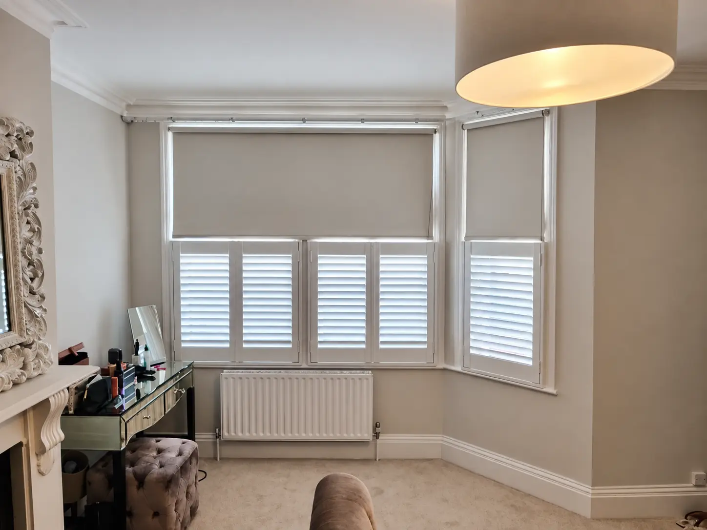 White cafe-style shutters across the lower part of a bedroom bay window, with roller blinds above.