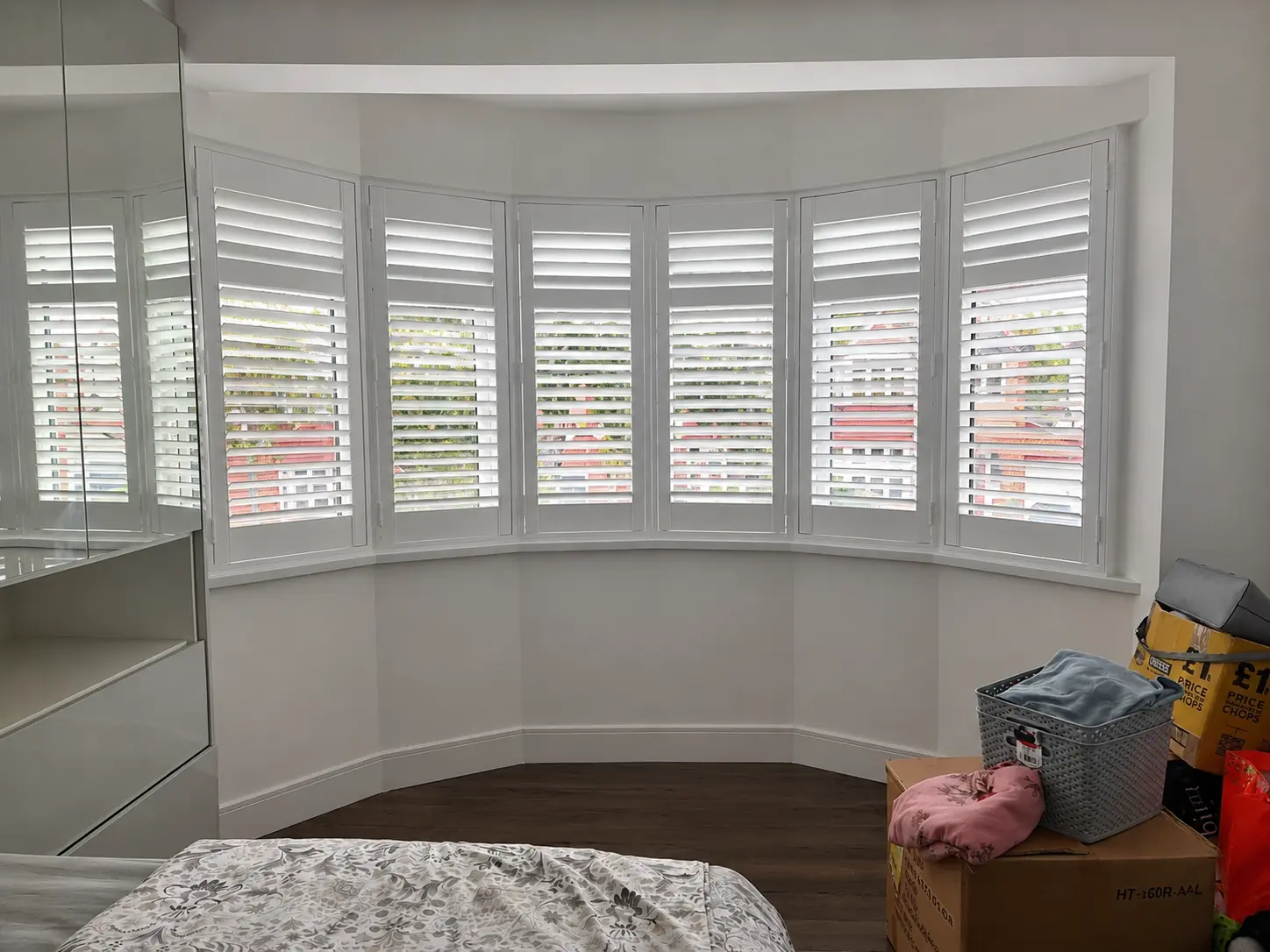 White full-height shutters following a wide, curved bedroom bay window beside mirrored wardrobes.