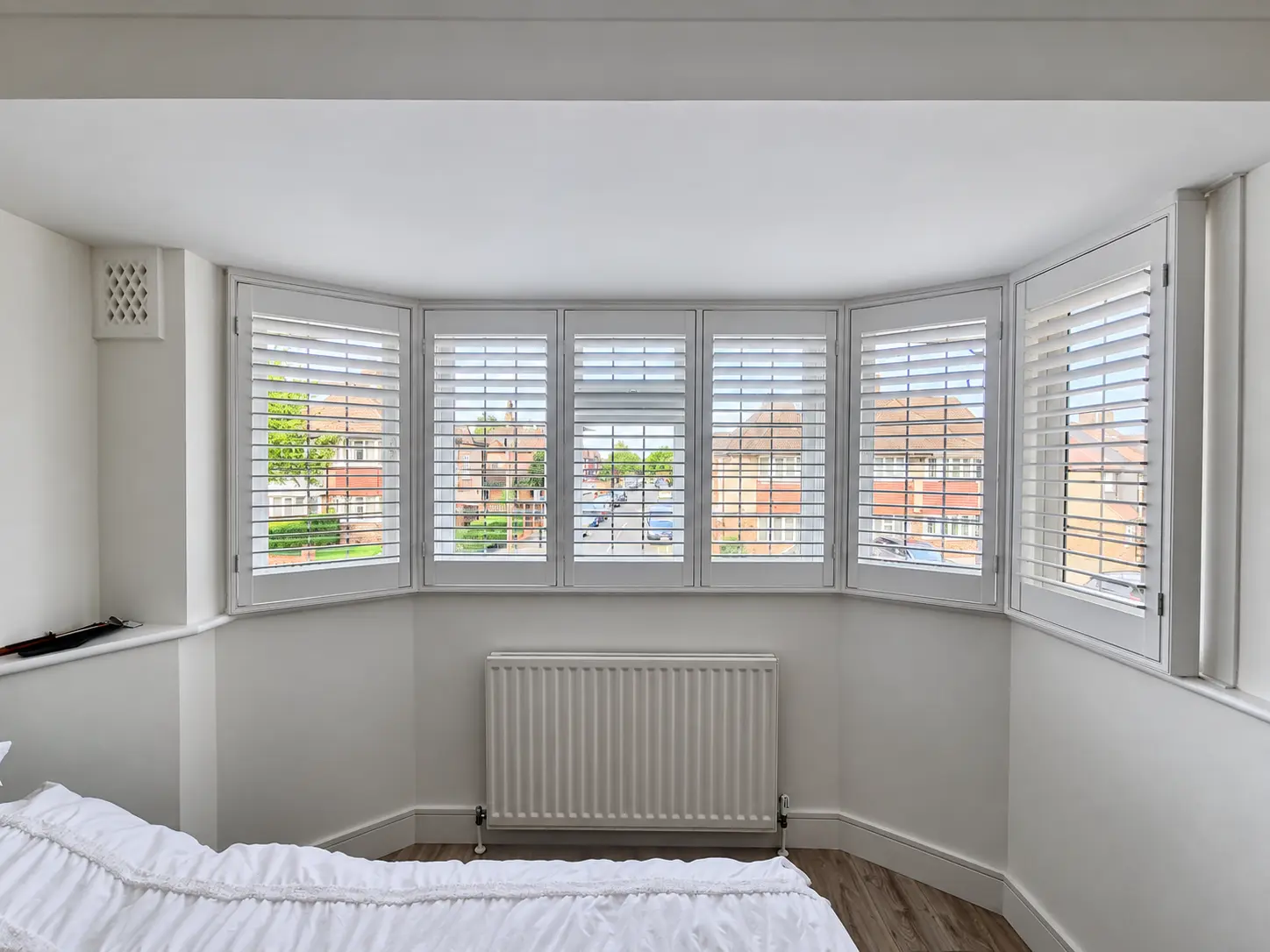White full-height shutters across a bedroom bay window above a radiator, with the louvres open to daylight.