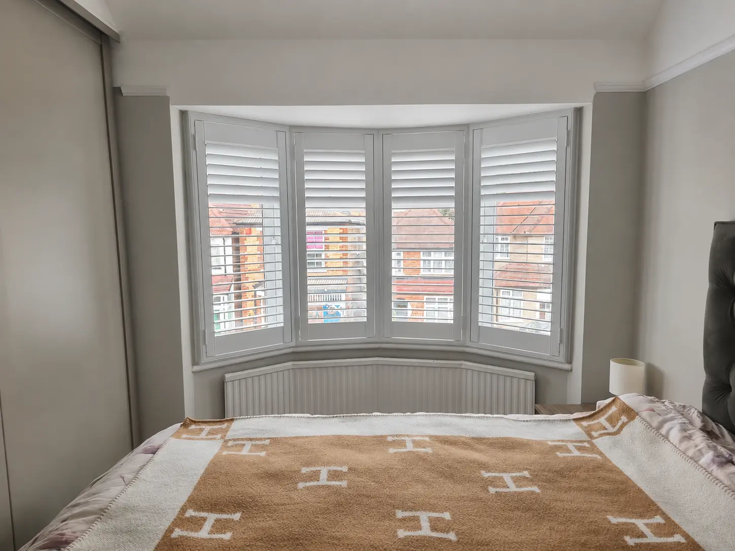 White full-height shutters in a bedroom bay window behind a bed, above a curved radiator.