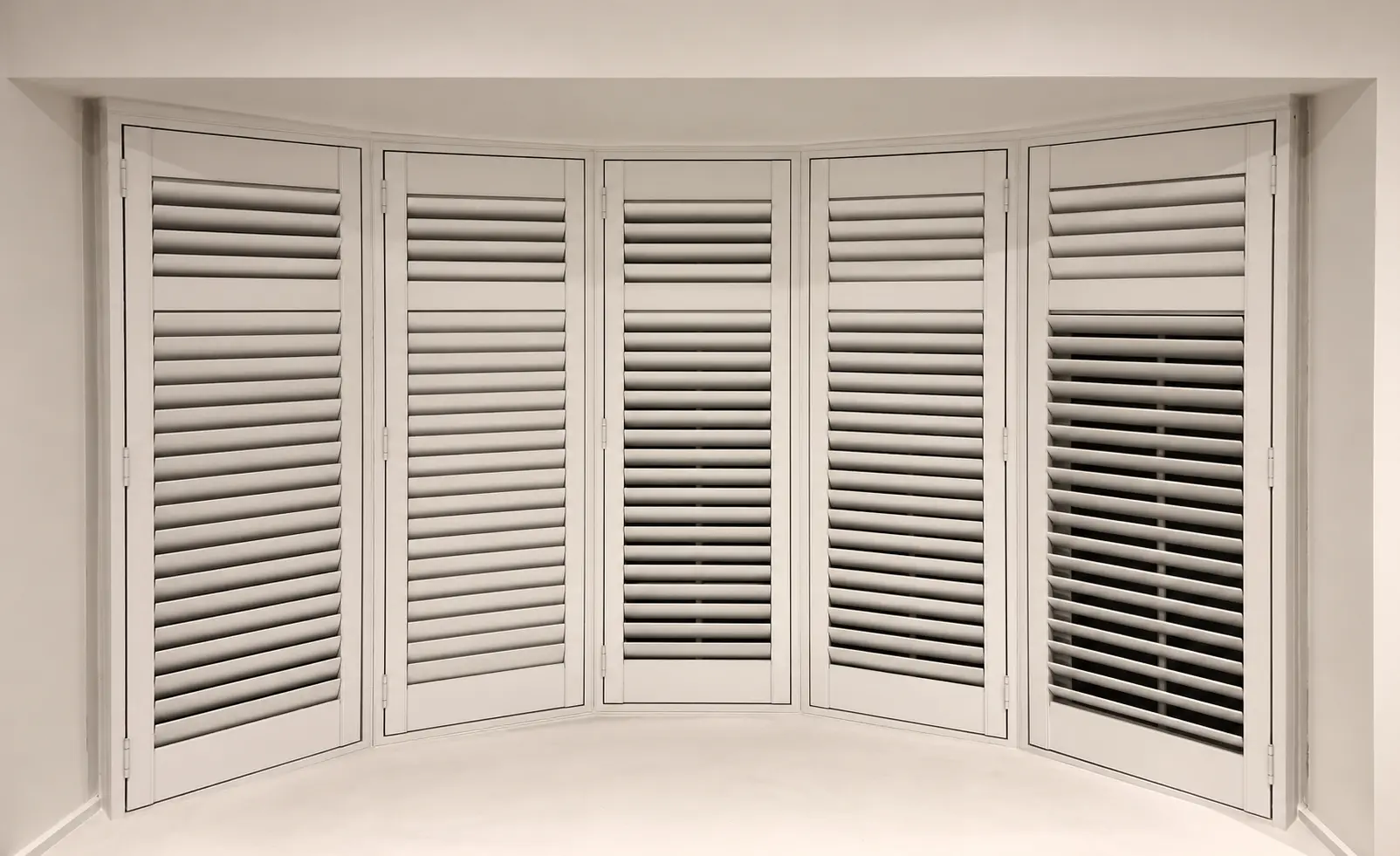 Five white full-height shutter panels following a living-room bay window, with the louvres closed.
