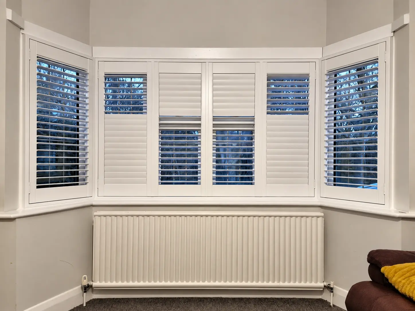 White full-height shutters across a living-room bay window above a radiator, with different louvre positions across the panels.