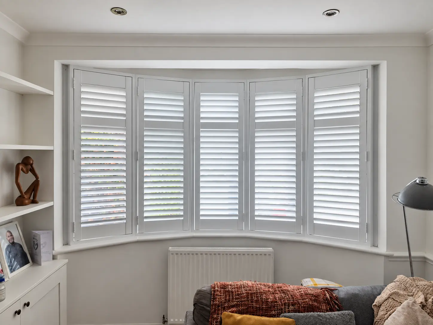 White full-height shutters in a living-room bay window beside built-in shelves and a sofa.