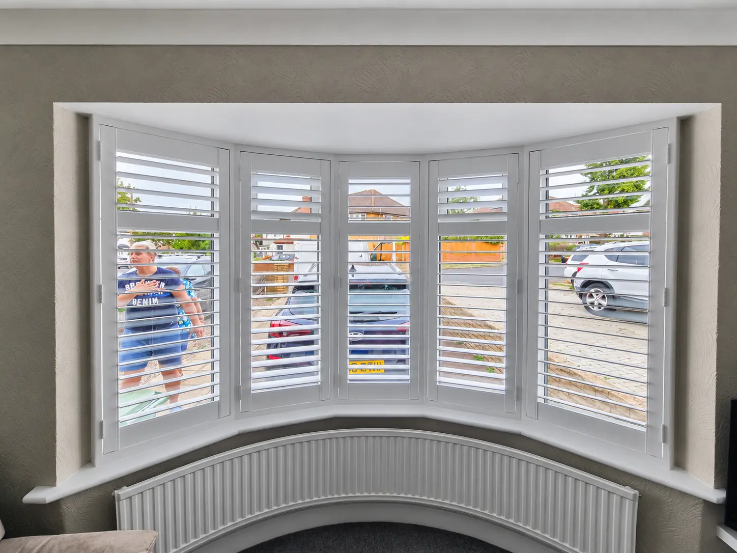 White full-height shutters following a curved living-room bay window above a curved radiator, with the louvres open.