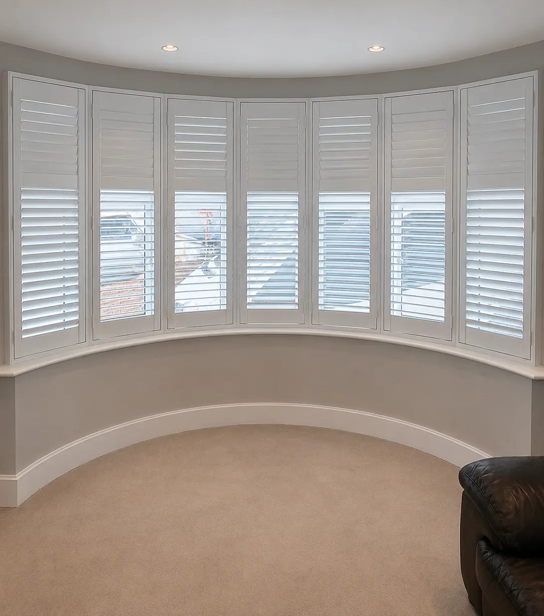 White full-height shutters following a broad, curved living-room bay window, with upper louvres closed and lower louvres open.