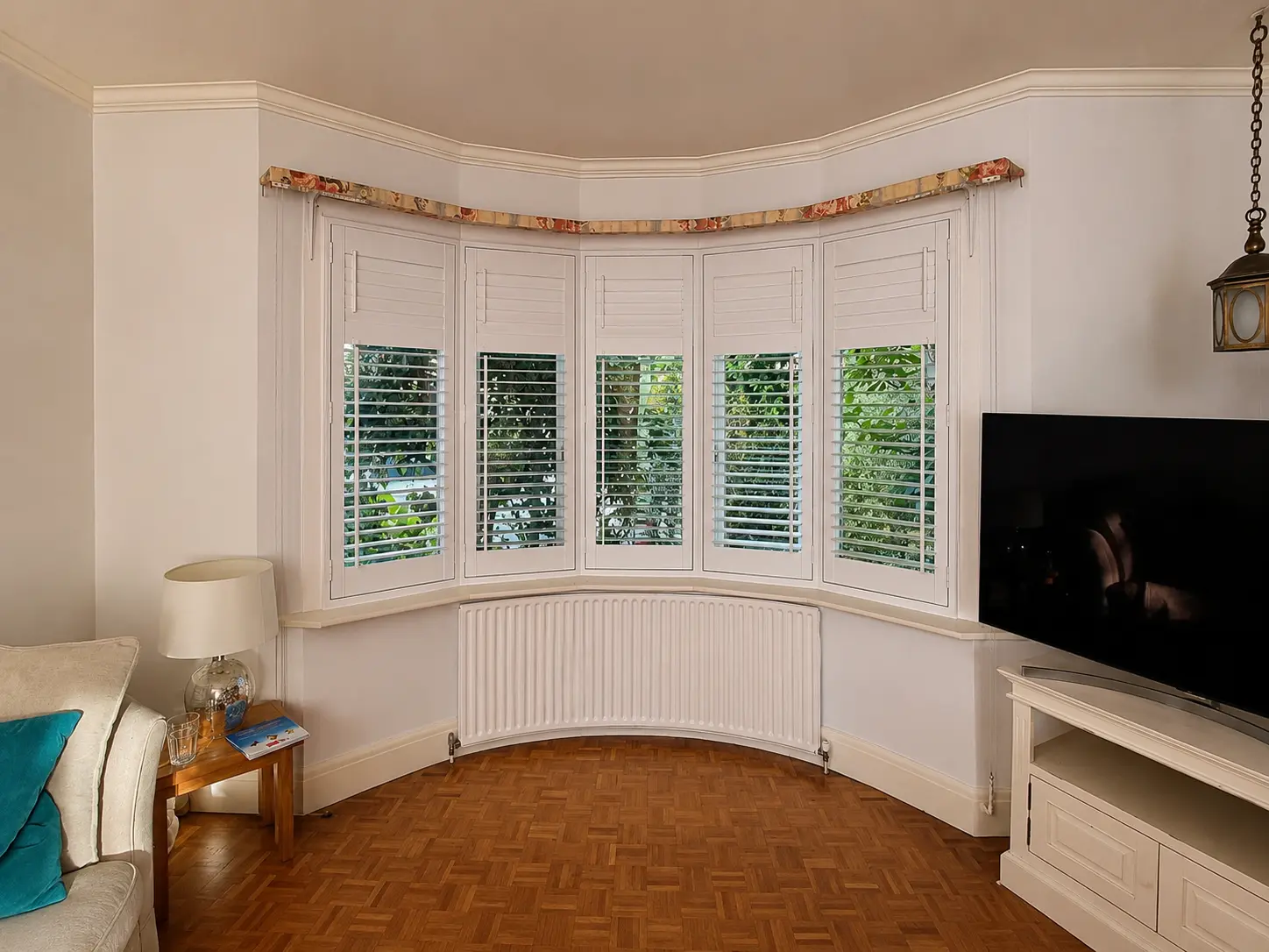 White full-height shutters in a curved living-room bay above parquet flooring, with upper louvres closed.