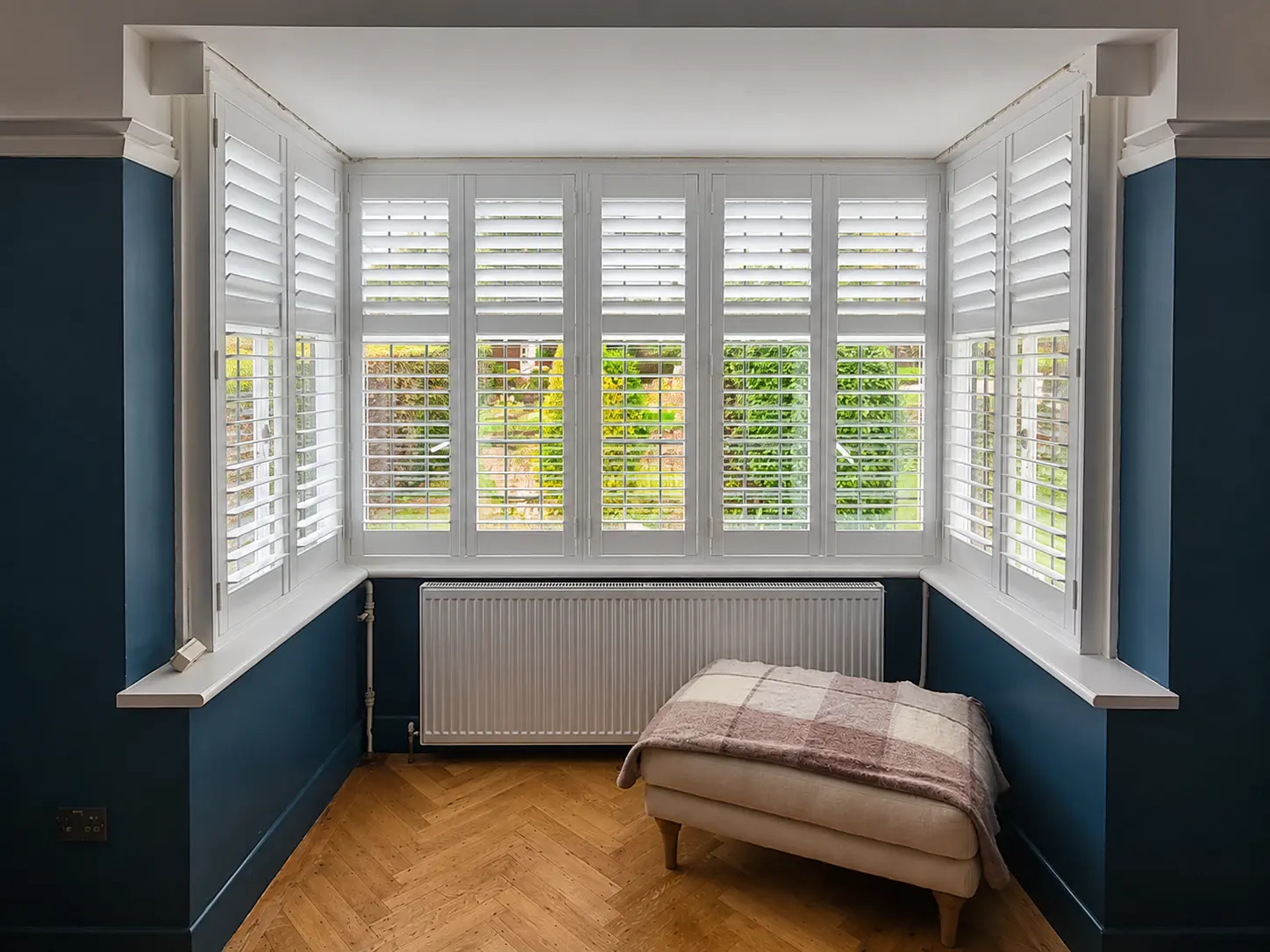 White full-height shutters with divider rails in a living-room bay with blue walls and a footstool.