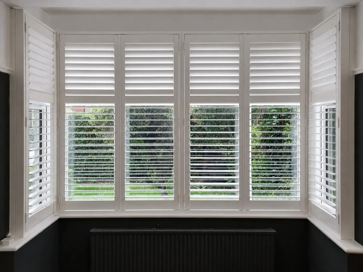 White full-height shutters across a living-room bay above a dark radiator, with upper louvres closed and lower louvres open.