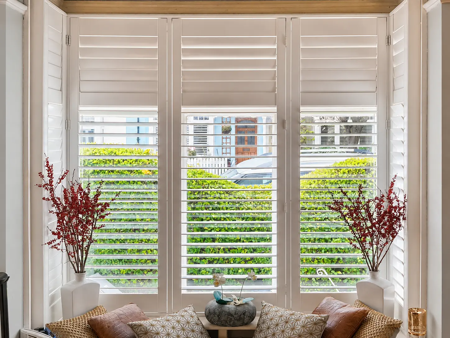 White full-height shutters across a tall living-room bay above a window seat, with upper louvres closed and lower louvres open.