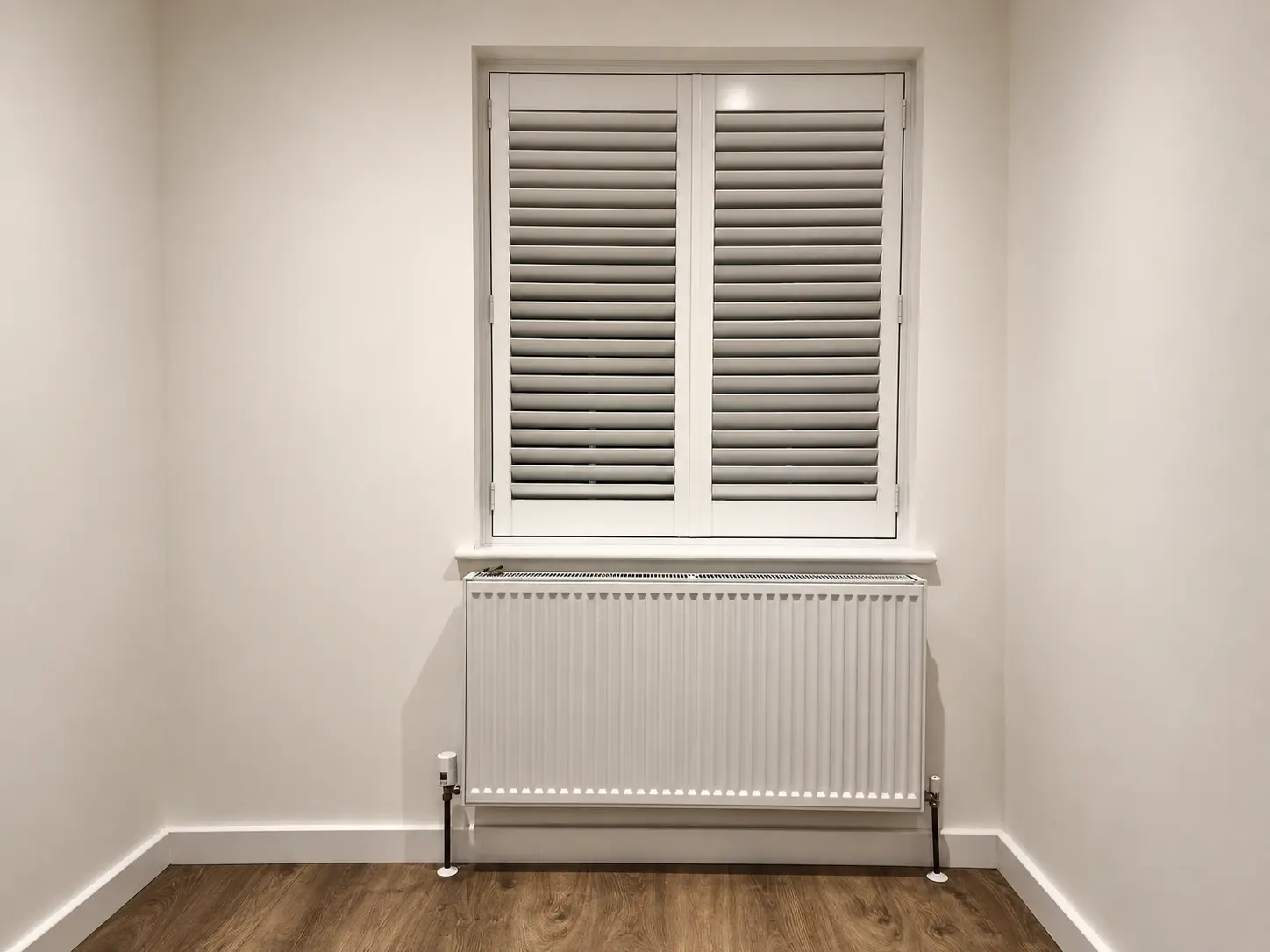 White full-height shutters at a bedroom window above a radiator in a plain white room.
