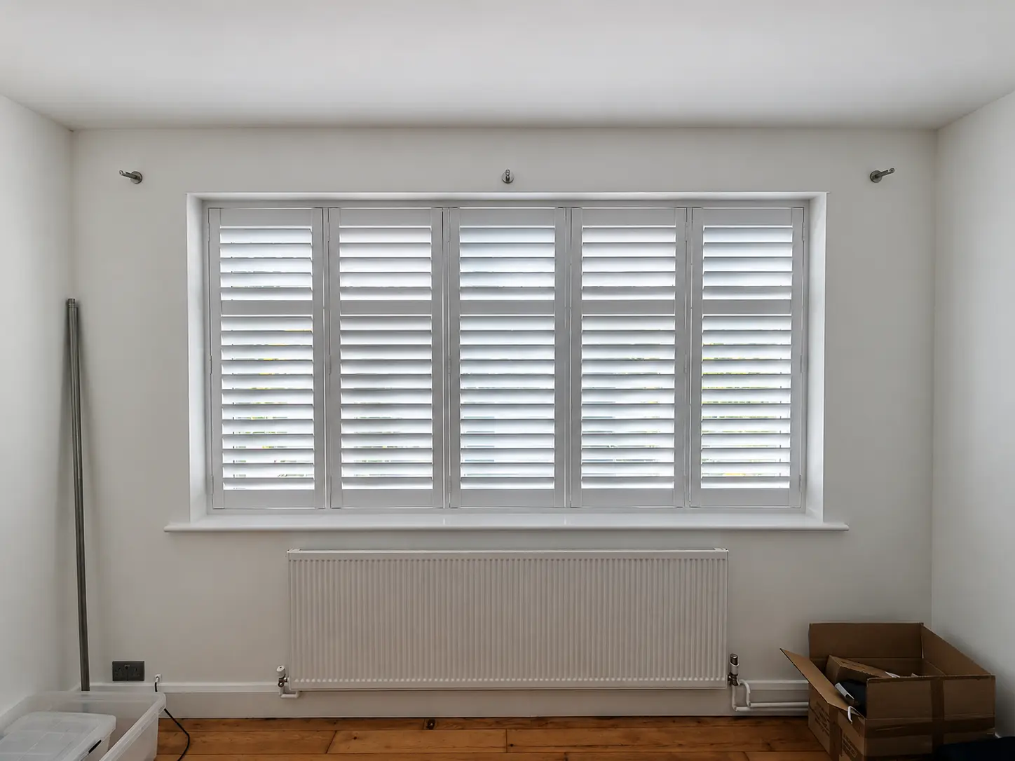 White full-height shutters spanning a wide bedroom window above a radiator.