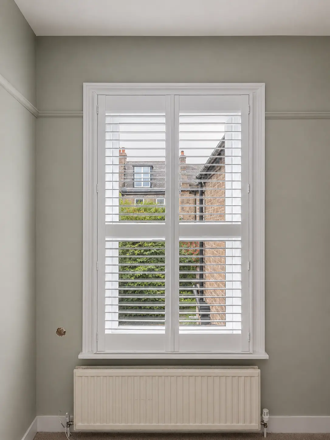 Two white full-height shutter panels with a divider rail at a tall bedroom window above a radiator.