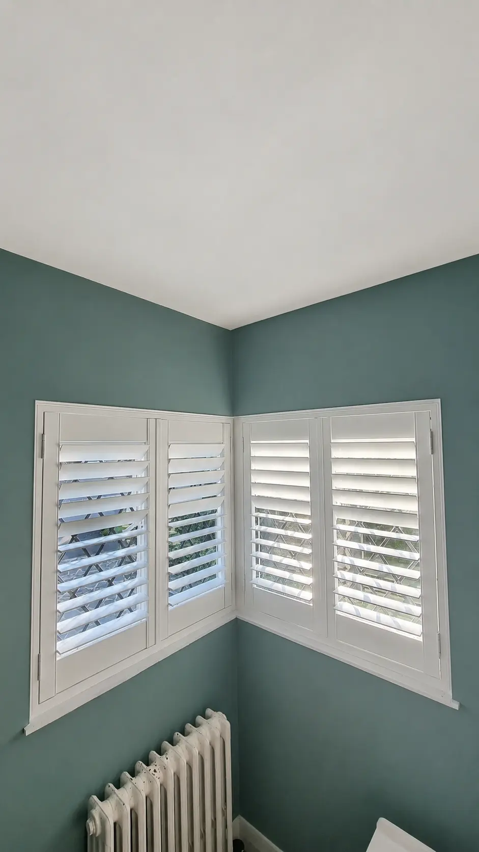 White full-height shutters fitted to two adjoining bedroom windows in a corner with teal walls.