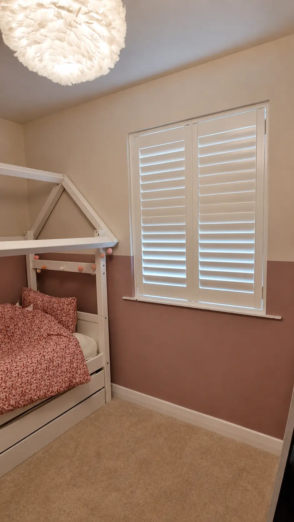 White full-height shutters at a bedroom window beside a house-frame bed and pink wall.