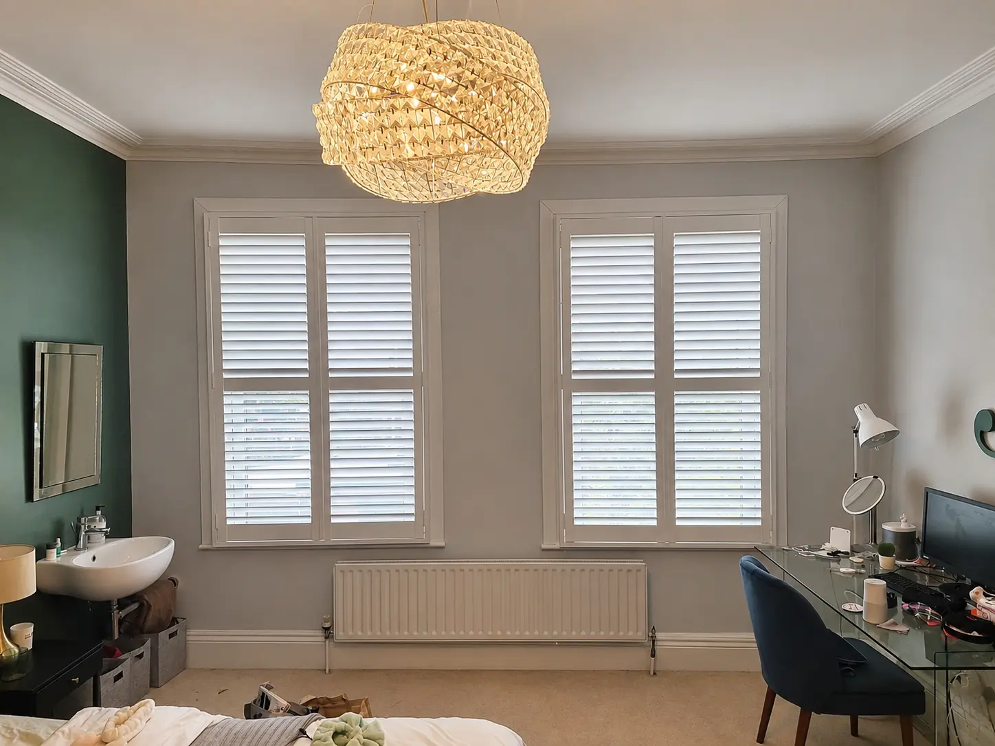 White full-height shutters with divider rails at two tall bedroom windows, one above a desk.