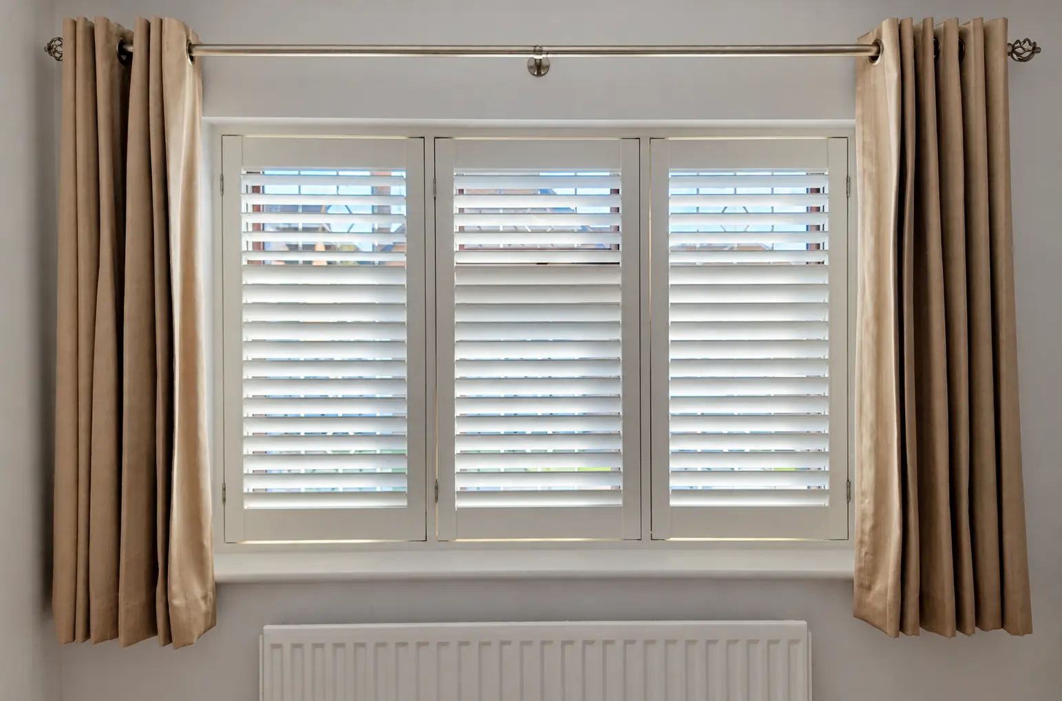 Three white full-height shutter panels at a dining-room window between beige curtains, with upper and lower louvres at different angles.