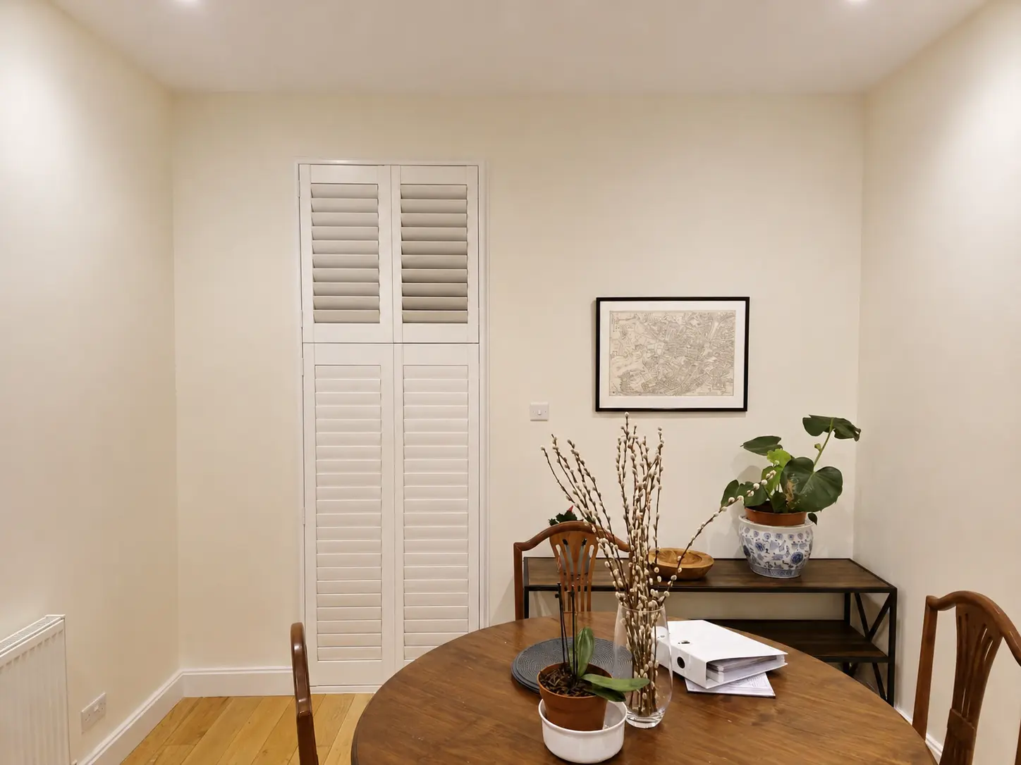 White full-height shutters with a divider rail at a tall, narrow window beside a dining table.