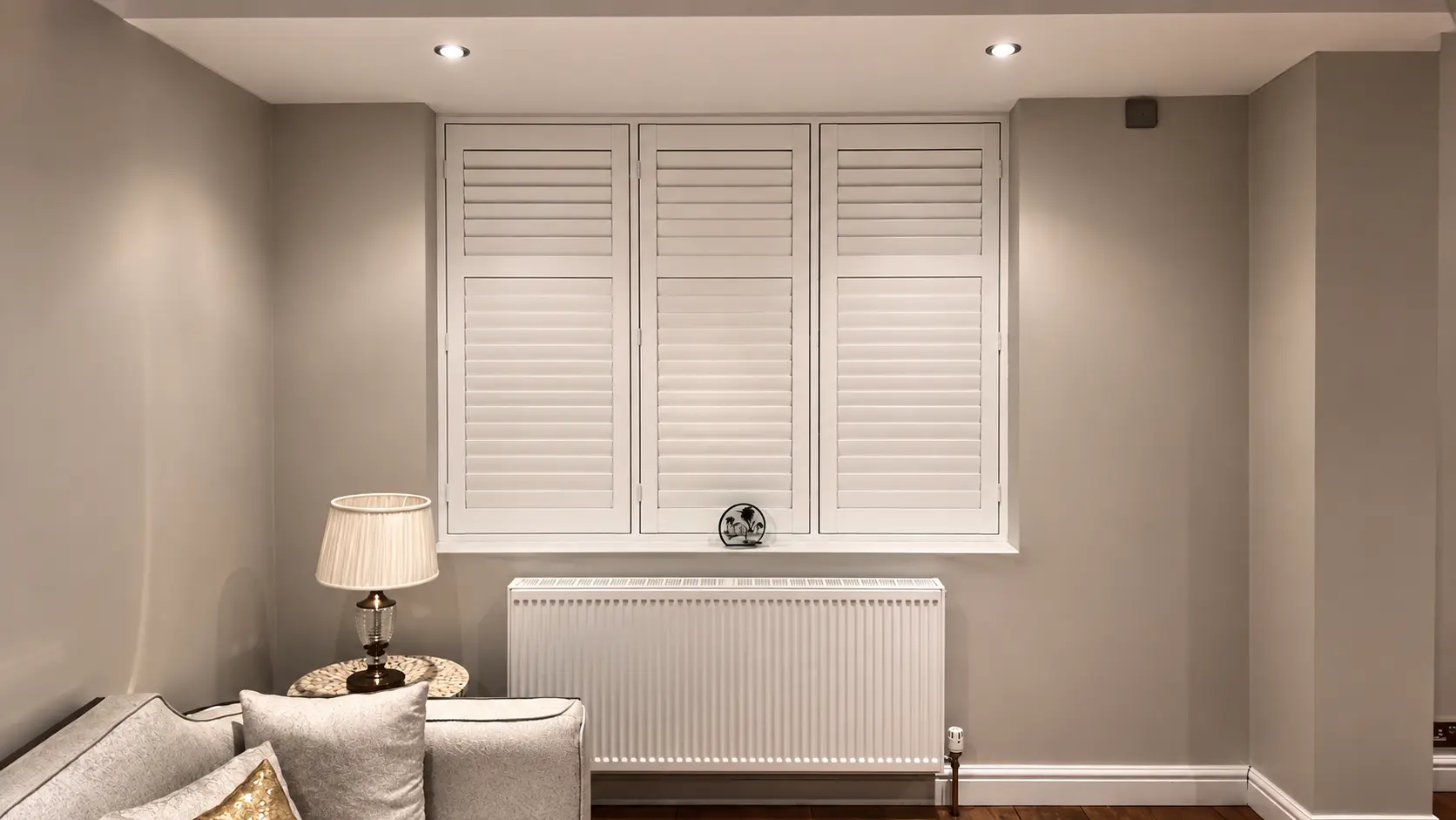 Three white full-height shutter panels with closed louvres above a radiator beside a sofa and table lamp.