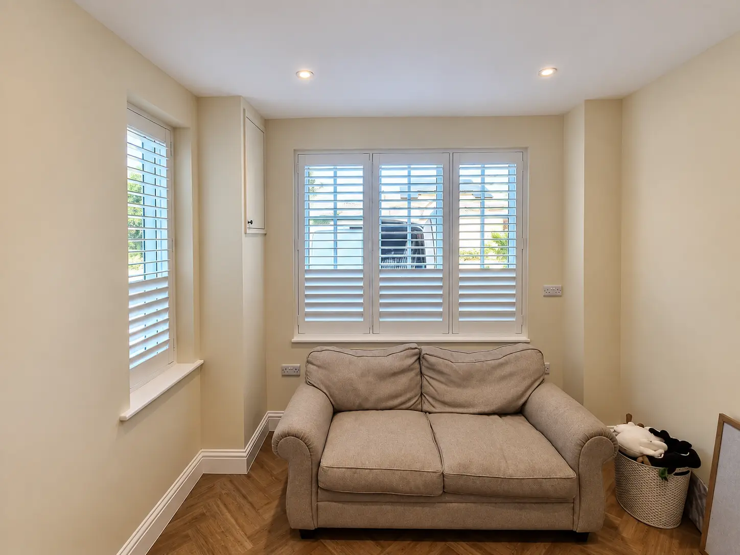 White full-height shutters at two living-room windows on adjoining walls beside a sofa.