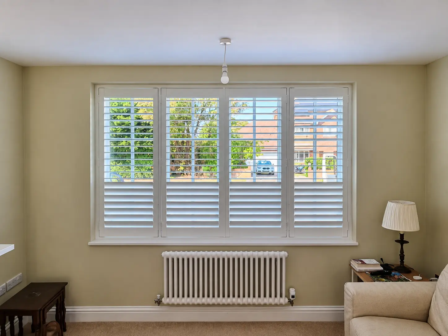 Four white full-height shutter panels across a living-room window above a radiator, with upper louvres open and lower louvres closed.