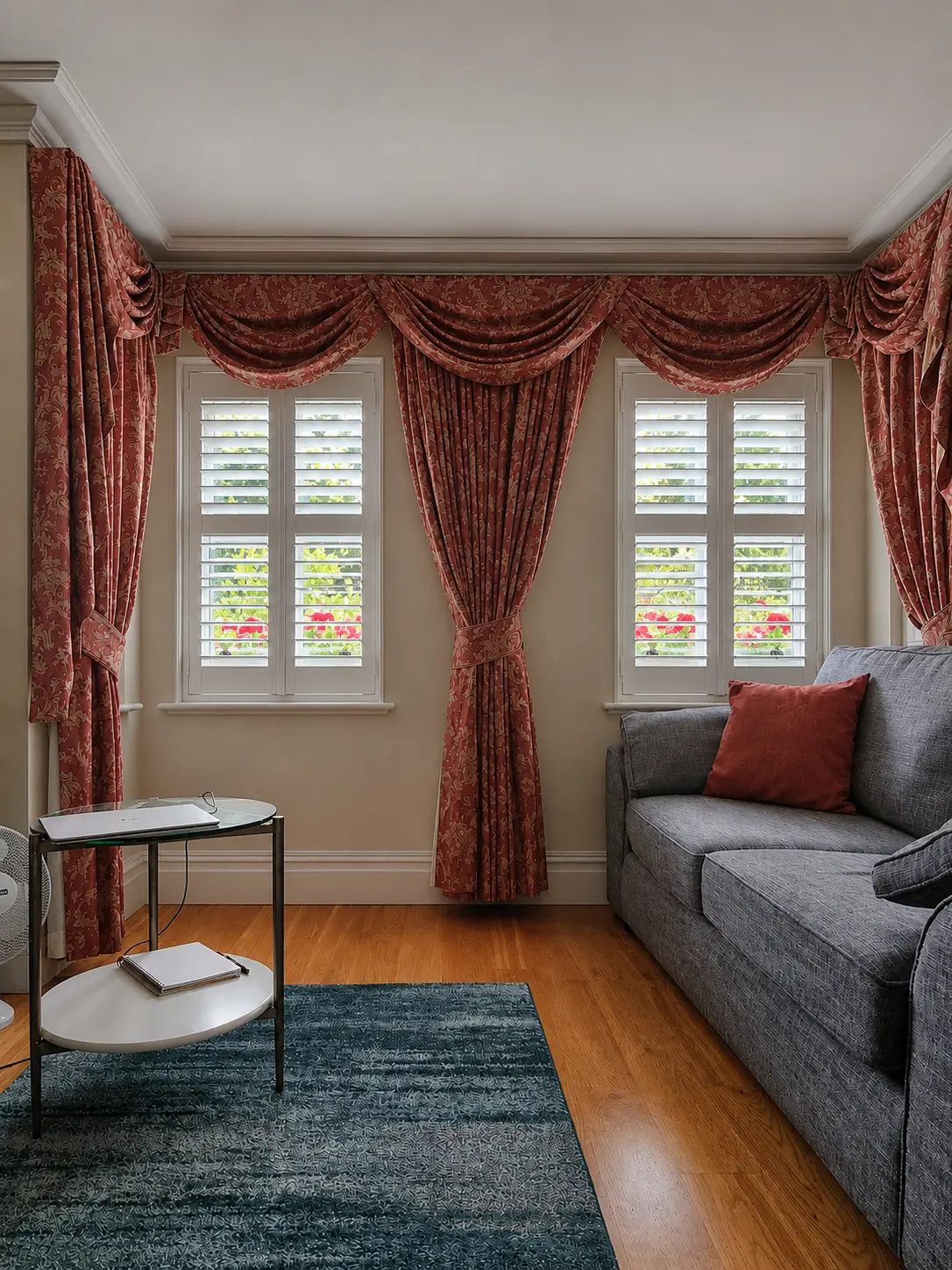 White full-height shutters at two living-room windows framed by red curtains beside a grey sofa.