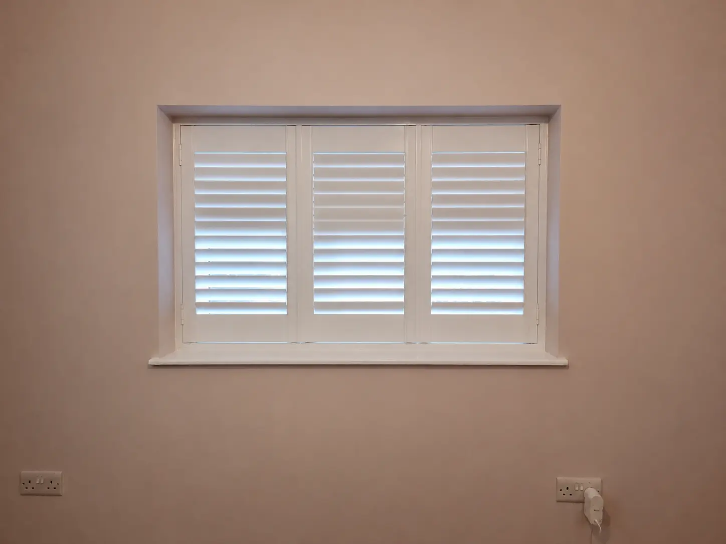 Three white full-height shutter panels at a living-room window in a pink wall, with upper louvres closed and lower louvres open.