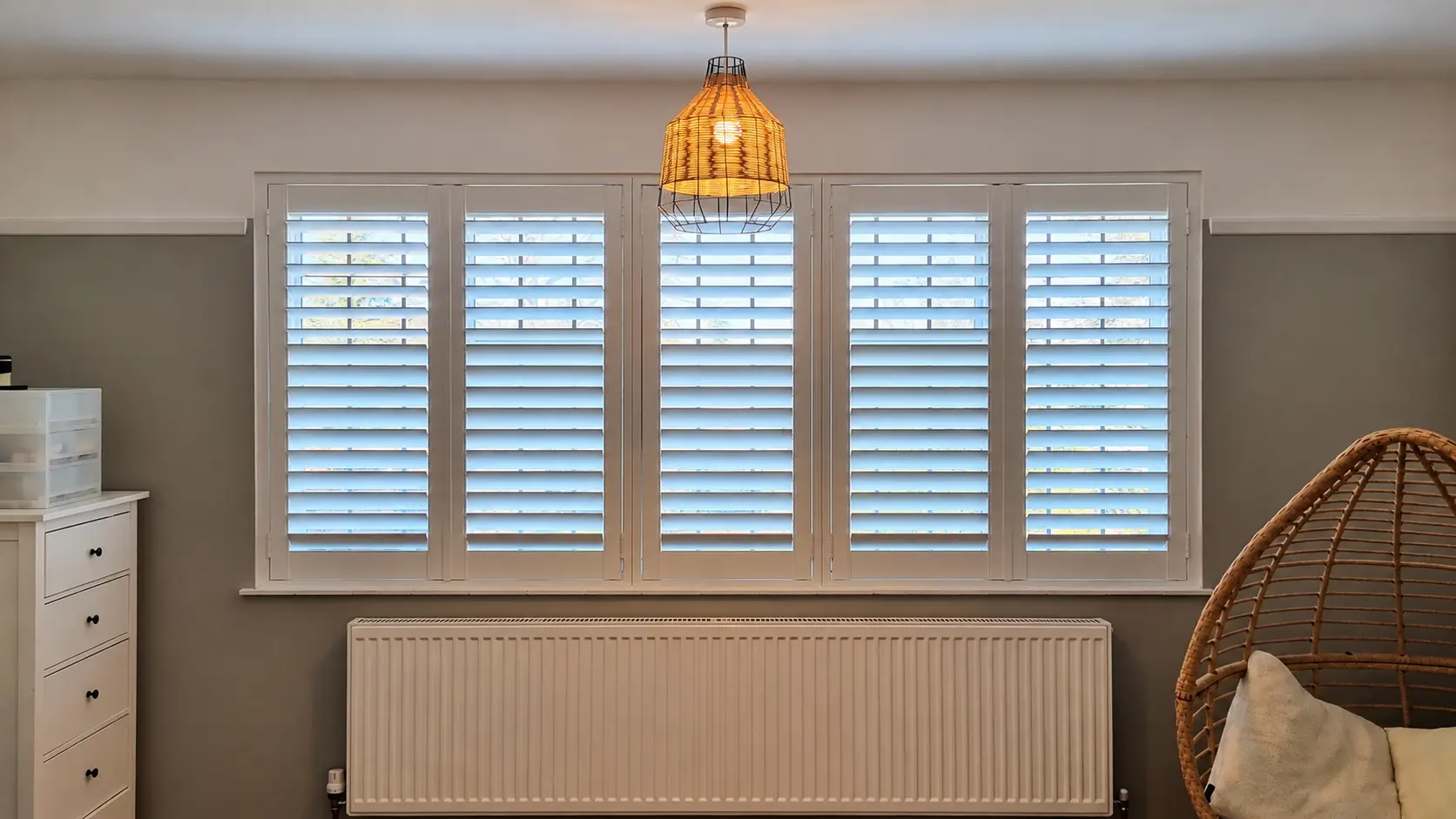 Five white full-height shutter panels across a living-room window above a radiator, beside a hanging chair.