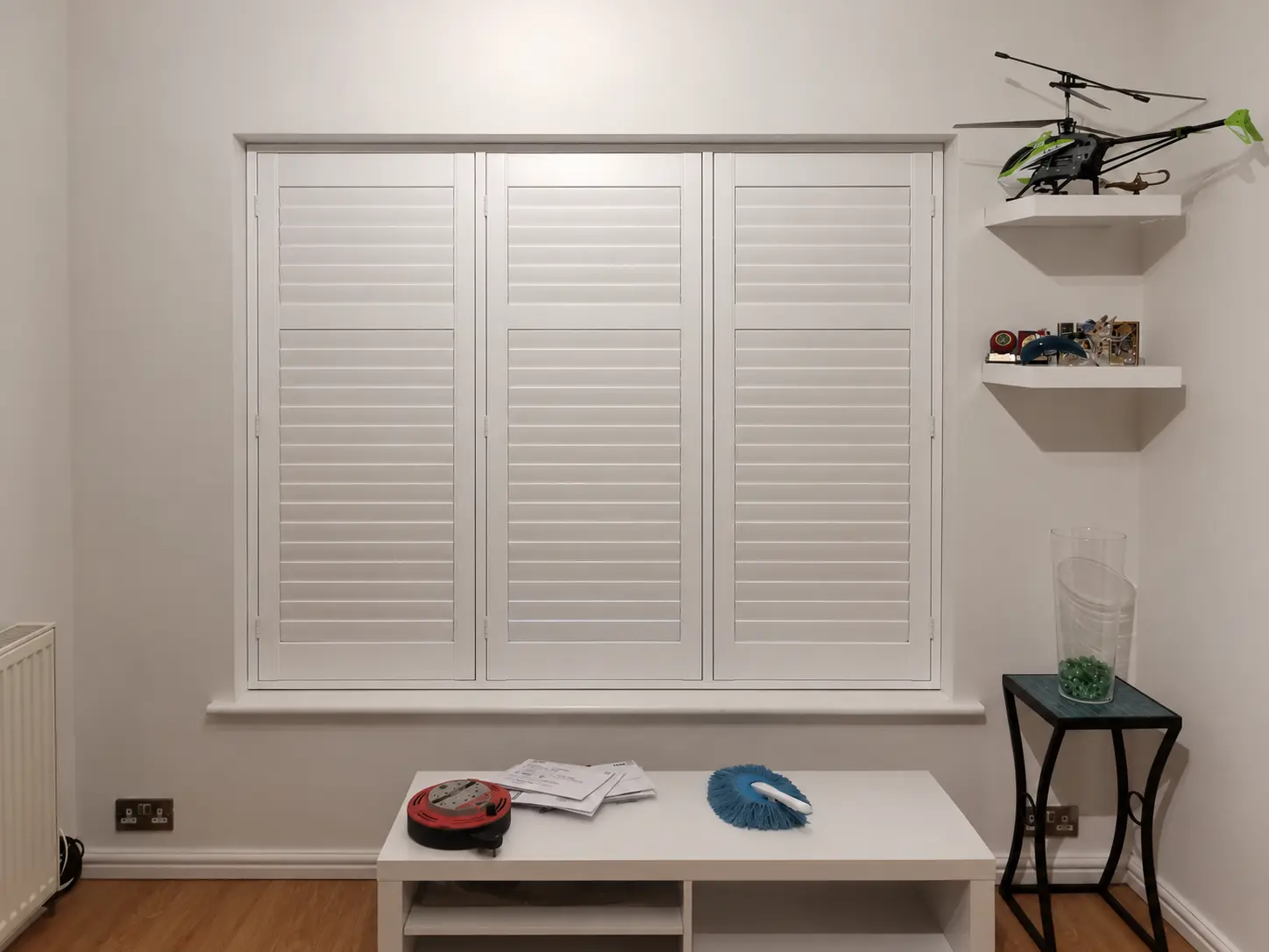 Three white full-height shutter panels with closed louvres at a window beside shelves displaying model aircraft.