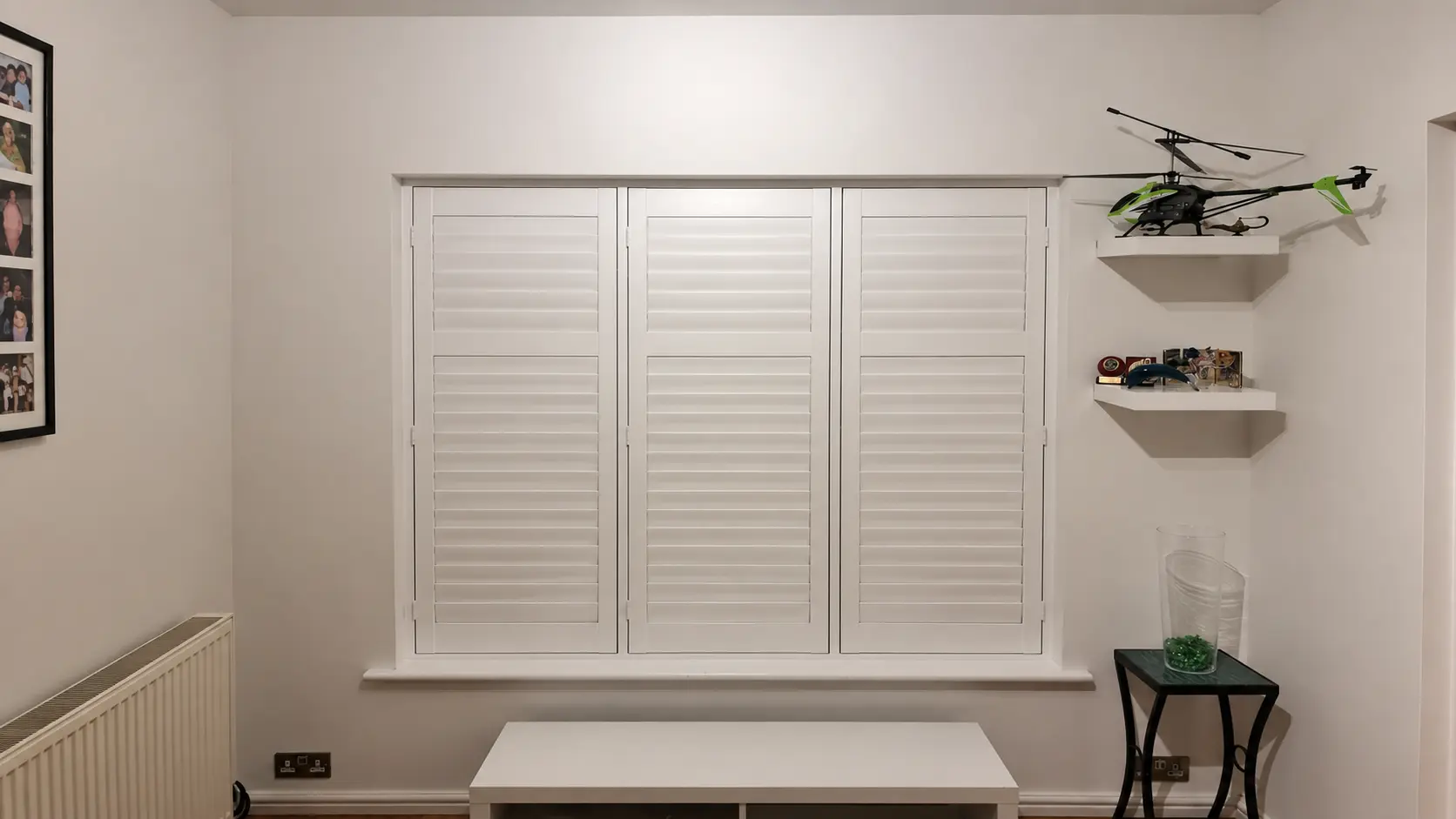 Three white full-height shutter panels with closed louvres at a window beside shelves displaying model aircraft, shown in a wider room view.