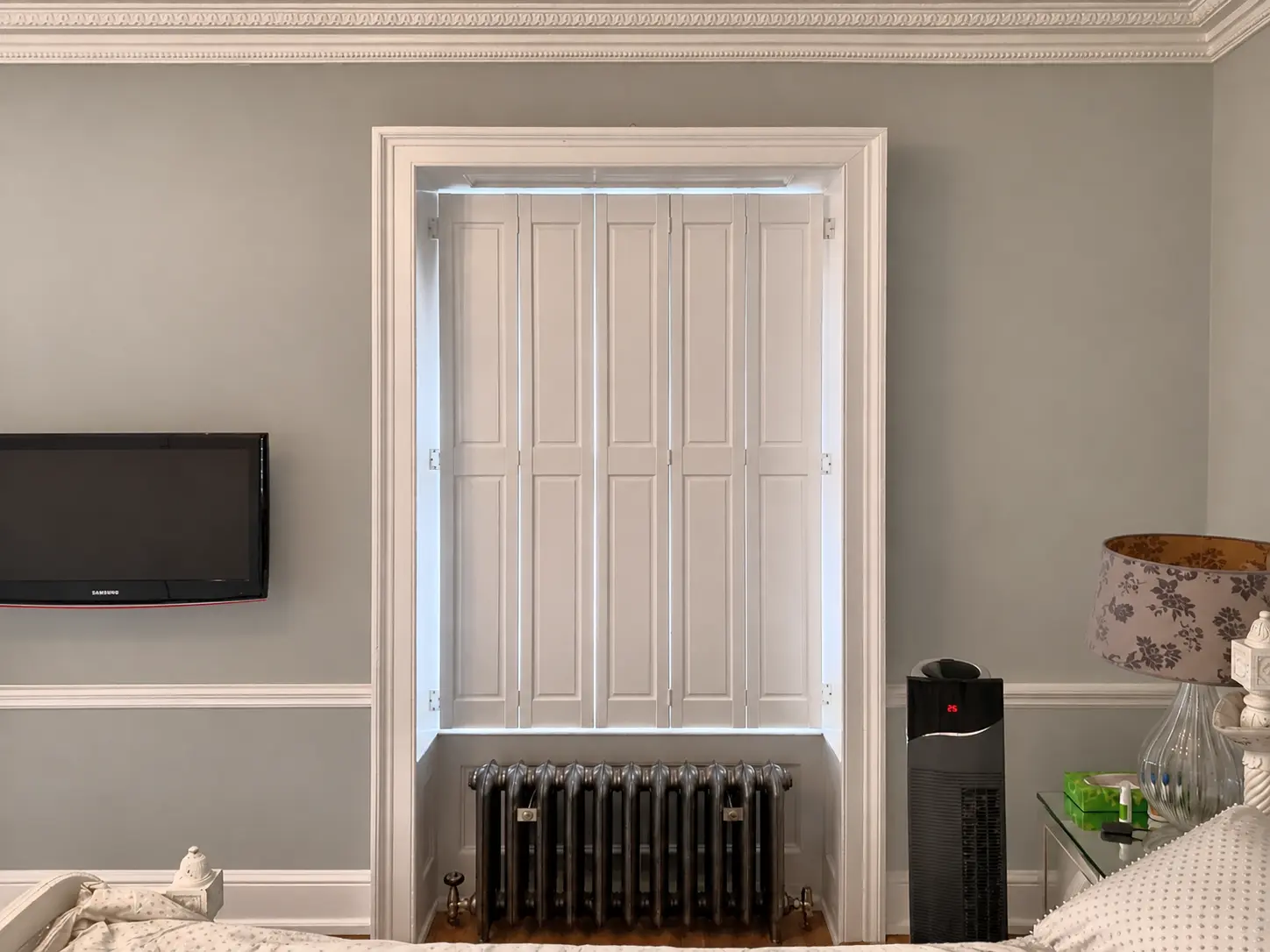 Closed white solid-panel shutters at a bedroom window above a black radiator, viewed from the left.