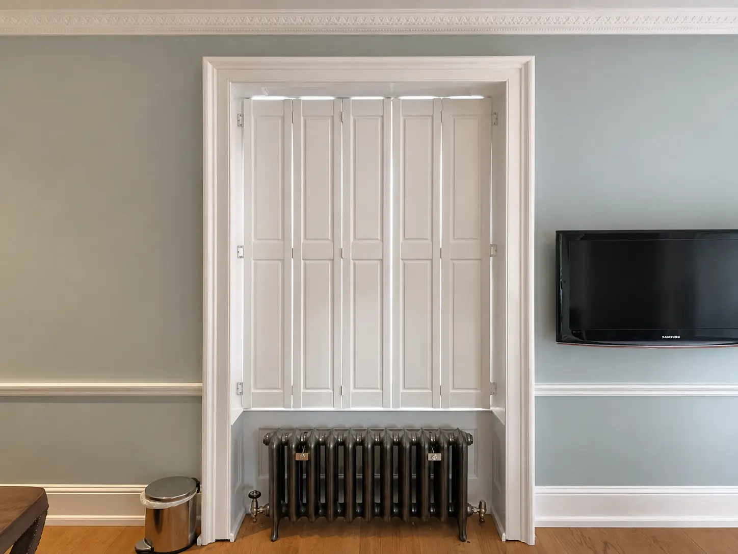 Closed white solid-panel shutters at a bedroom window above a black radiator, viewed from the right.