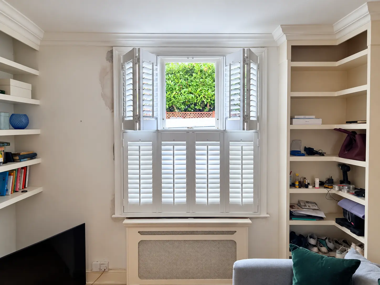 White tier-on-tier shutters at a living-room window, with the upper panels folded open and lower panels closed.